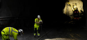 Two surveyors work in a dark rail tunnel in front of excavation equipment 