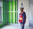 A construction professional wearing a red safety vest and a white hard hat equipped with a 360-degree reality capture camera stands inside an unfinished building. The room features exposed metal framing with green insulation panels, a concrete wall, and a window allowing natural light. The individual is smiling, representing the use of cutting-edge digital solutions for construction progress tracking. The image highlights Hexagon’s Building Solutions "Create" portfolio, showcasing the role of advanced reality capture technology in digital construction workflows.