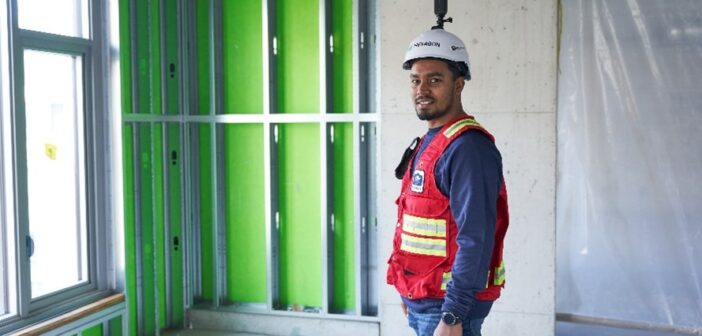 A construction professional wearing a red safety vest and a white hard hat equipped with a 360-degree reality capture camera stands inside an unfinished building. The room features exposed metal framing with green insulation panels, a concrete wall, and a window allowing natural light. The individual is smiling, representing the use of cutting-edge digital solutions for construction progress tracking. The image highlights Hexagon’s Building Solutions "Create" portfolio, showcasing the role of advanced reality capture technology in digital construction workflows.