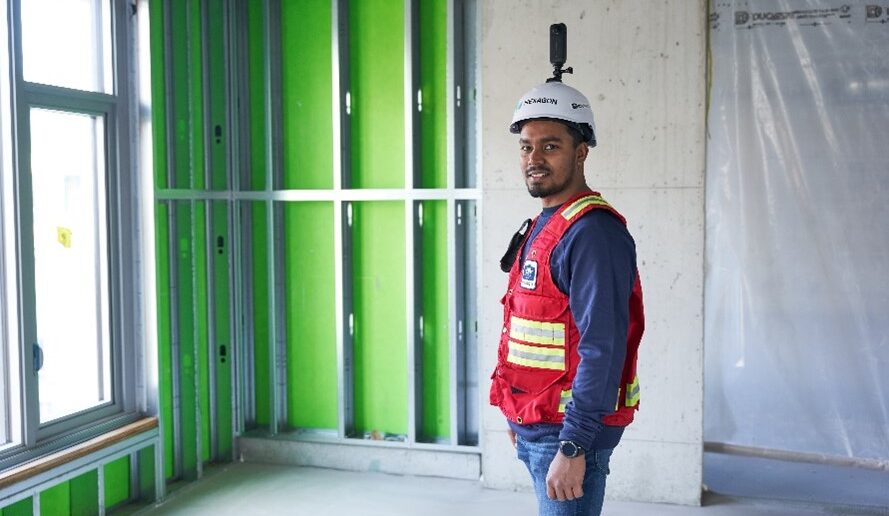 Caption: "Reality Capture in Action: Enhancing Construction Progress Tracking with Hexagon’s Building Solutions" Alt Text: A construction professional wearing a red safety vest and a white hard hat equipped with a 360-degree reality capture camera stands inside an unfinished building. The room features exposed metal framing with green insulation panels, a concrete wall, and a window allowing natural light. The individual is smiling, representing the use of cutting-edge digital solutions for construction progress tracking. The image highlights Hexagon’s Building Solutions "Create" portfolio, showcasing the role of advanced reality capture technology in digital construction workflows.